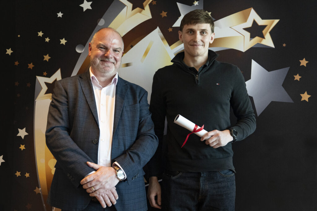 A smiling man in a suit standing next to a taller smiling young man in a black shirt, who holds a rolled-up certificate. They are against a star-patterned background.