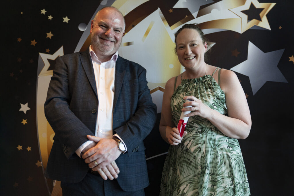  A smiling man in a suit and a smiling woman in a floral dress holding a certificate, against a star-patterned background.