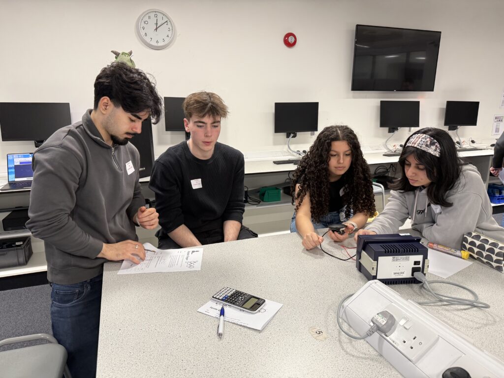 Four individuals are gathered around a gray laboratory table in a classroom setting. The table has a calculator, pen, and paper on it, along with an electronic device connected to wires. The background shows several computer monitors mounted on the wall, a clock, and a power strip on the table. The group appears to be engaged in a hands-on activity involving the equipment.